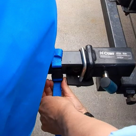 Close-up of blue nylon strap being fastened to trailer hitch to secure Riptide cover during transport